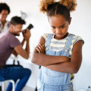 girl with crossed arms in foreground, parents blurry in back